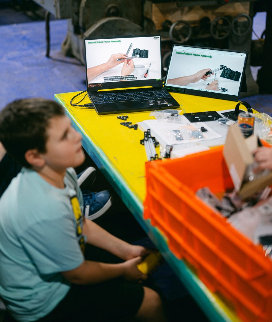 A BeanBots student watches a REV Robotics frame-assembly video at a worktable full of robot parts